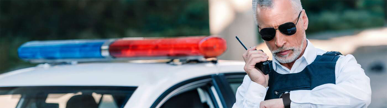 Police officer on a walkie talkie standing in front of a police car.