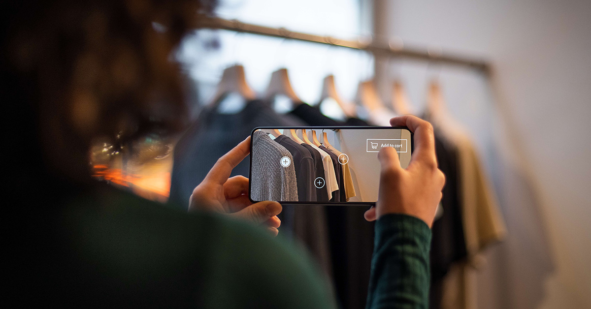 A shopper uses a smartphone with an AI-powered app to scan and add clothing items to a cart in a retail store.